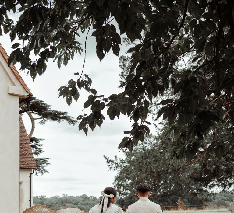 Bride in long mesh sleeve spotted Vagabond wedding dress with long train and bridal hair bow walks into churchyard holding hands with groom linen suit and brown shoes at Wasing Park wedding