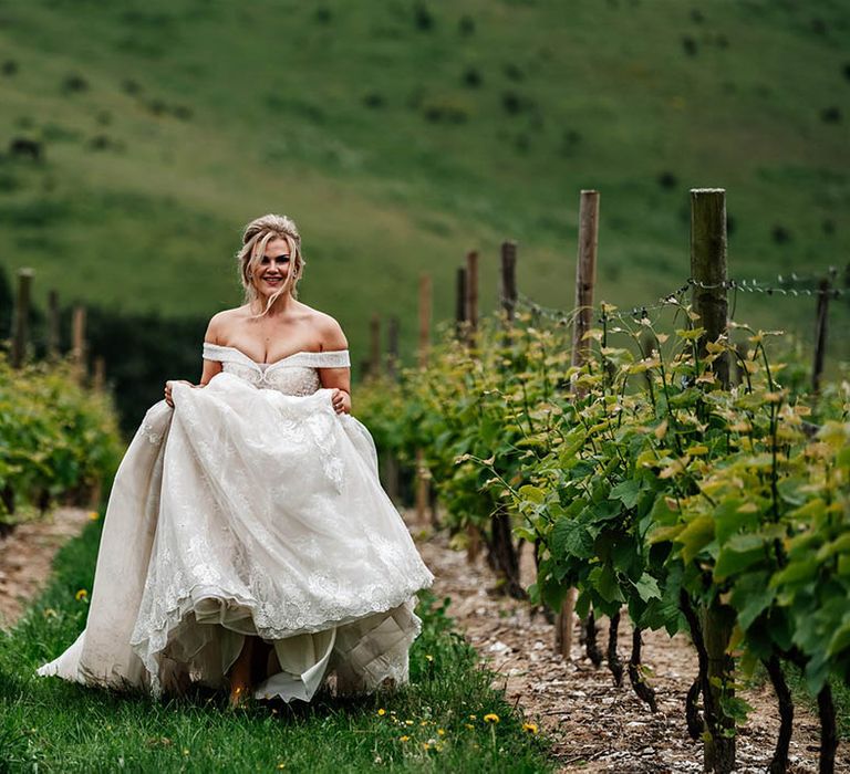 Bride in an off the shoulder princess wedding dress running through Little Wold Vineyard