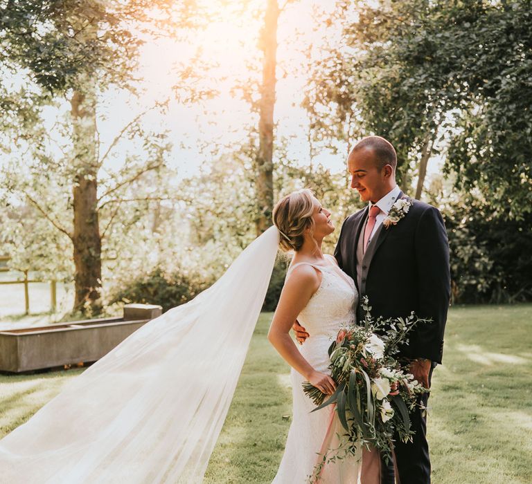 Bride in white lace Justin Alexander wedding dress with train and veil holding white, pink and green bridal bouquet stands with groom in dark three piece suit with pink tie in the gardens at Tythe Barn wedding with barn wedding flowers