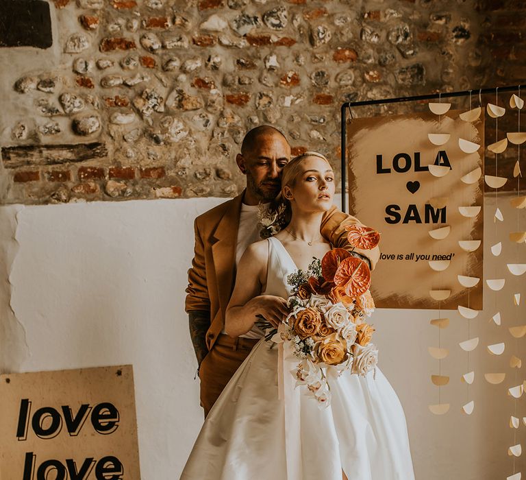 Groom in a mustard suit embracing his bride in a Jesus Peiro wedding dress with front split as she holds a tropical wedding bouquet with orange anthuriums 