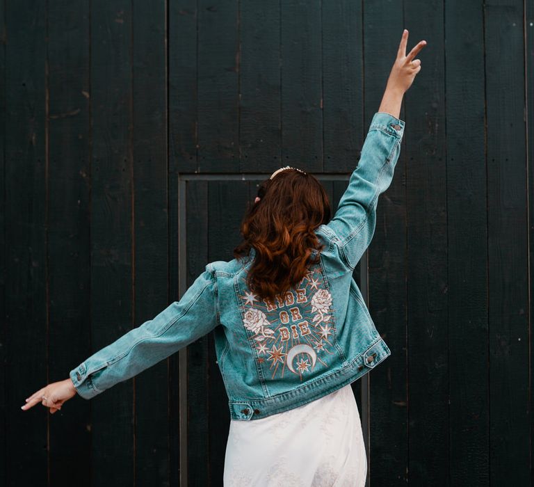 Bride in a denim jacket with gold Ride Or Die embroidered design on the back holding her bouquet in the air 