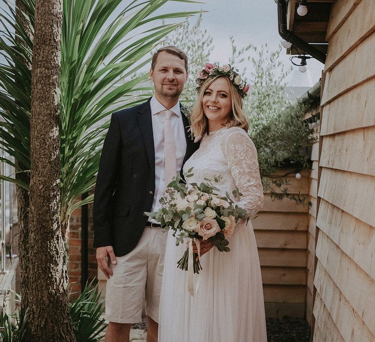 Bride and groom portrait at back garden wedding with groom in beige shorts and a navy blazer and the pregnant bride in a floaty wedding dress with long sleeves 