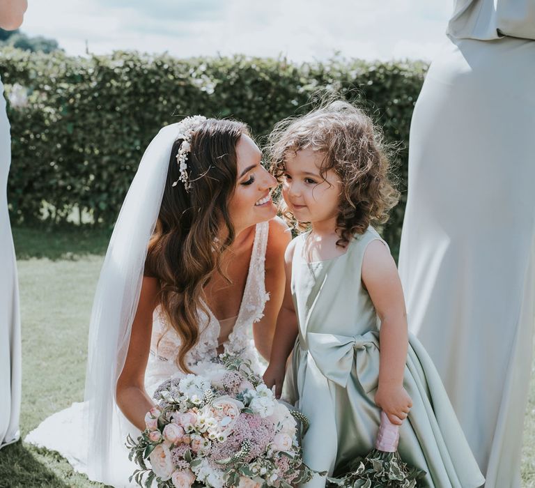 Bride with curled hair wearing Berta wedding dress, headband and veils holding white, pink and green bridal bouquet smiles with flower girl in sage green satin dress for summer wedding at Primrose Hill Farm