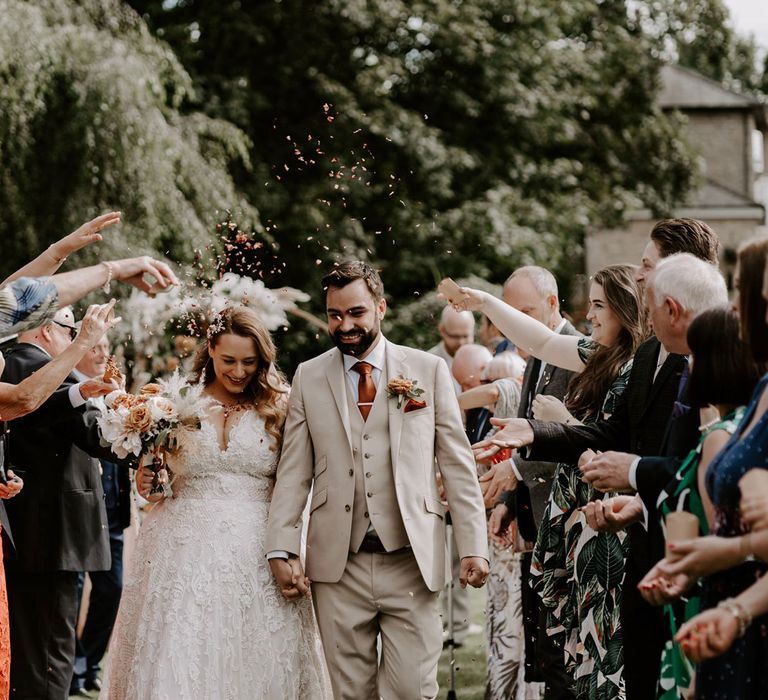 Bride in homemade lace wedding dress and veil holding rose bridal bouquet walks holding hands with groom in beige three piece suit whilst guests throw confetti during DIY garden wedding ceremony