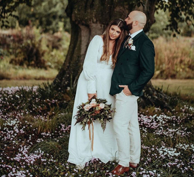 Bride leans into groom on their wedding day as they stand beside tree