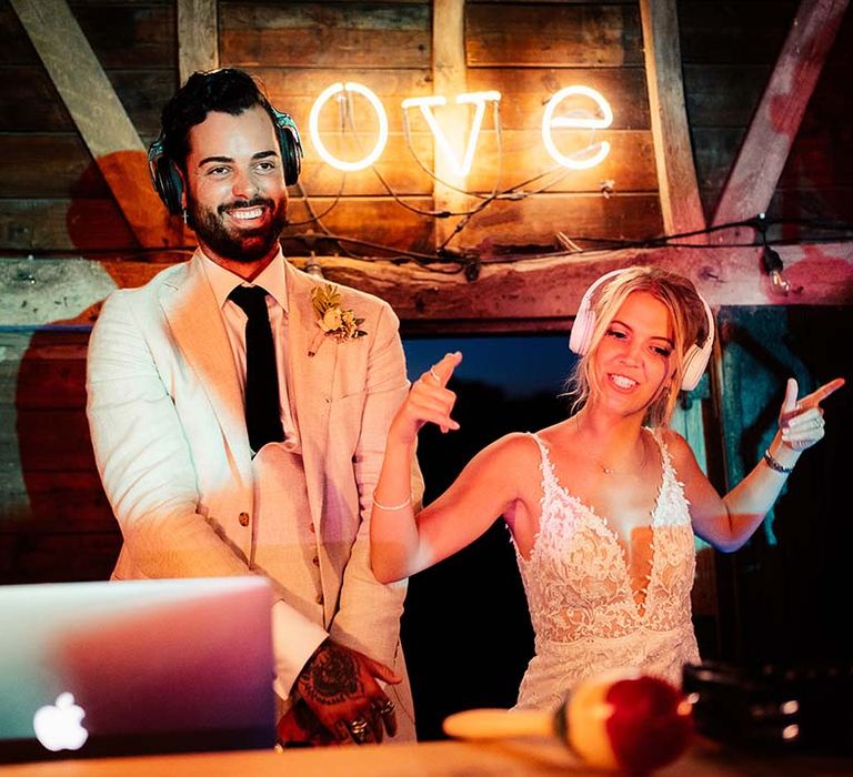 Groom in a beige suit and black tie in the DJ booth with his bride in a lace dress and headphones 