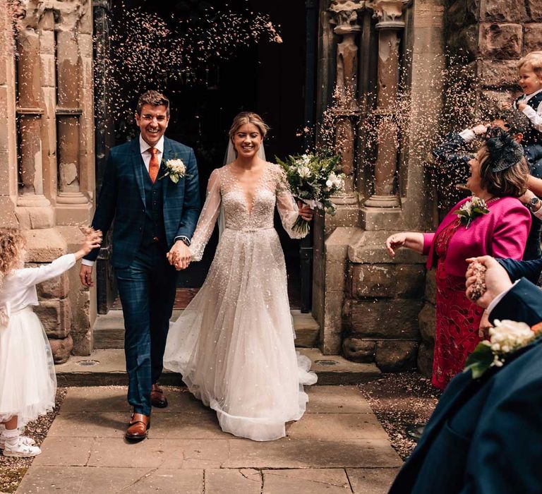 Groom in blue checked three piece suit and tie walks out of church with bride in long sleeve embroidered wedding dress and veil as guests throw confetti