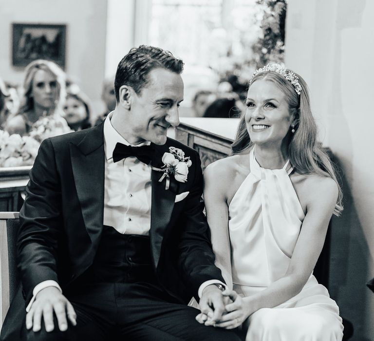 Smiling bride in halterneck Halfpenny London wedding dress with bespoke floral headband sits with groom in black tie and floral buttonhole during church wedding ceremony