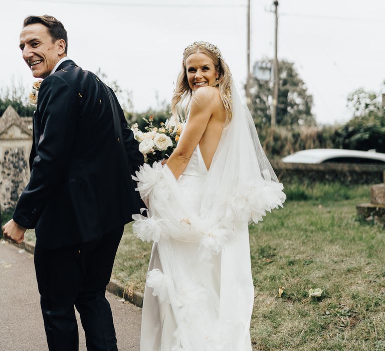 Bride in Halfpenny London wedding dress and veil with floral headband holds hands with groom in black tie as they walk out of churchyard after wedding ceremony