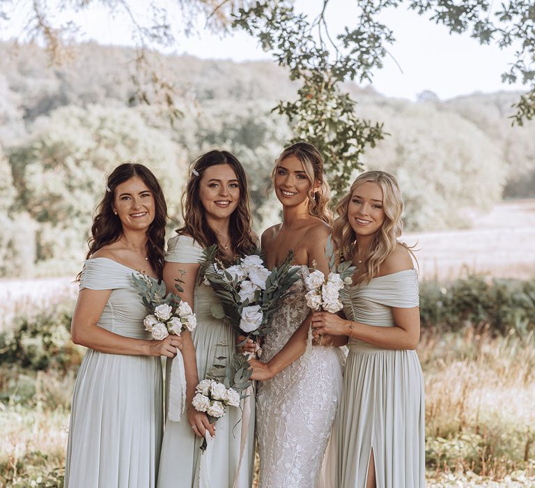 Bride stands with her bridesmaids outdoors who hold floral bouquets