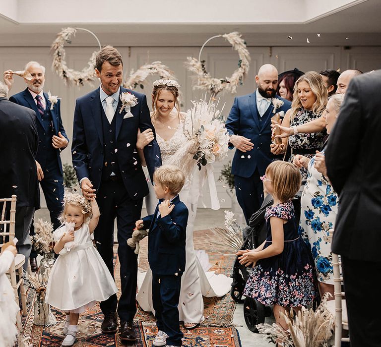 Bride & groom walk down the aisle with their children as wedding guests throw confetti around them