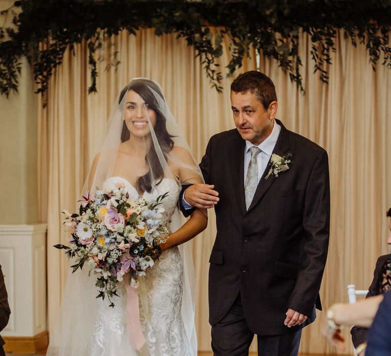 Bride in lace and tulle wedding dress with veil over her head holding pastel bridal bouquet walks down the aisle arm in arm with her father in a dark suit white white rose buttonhole