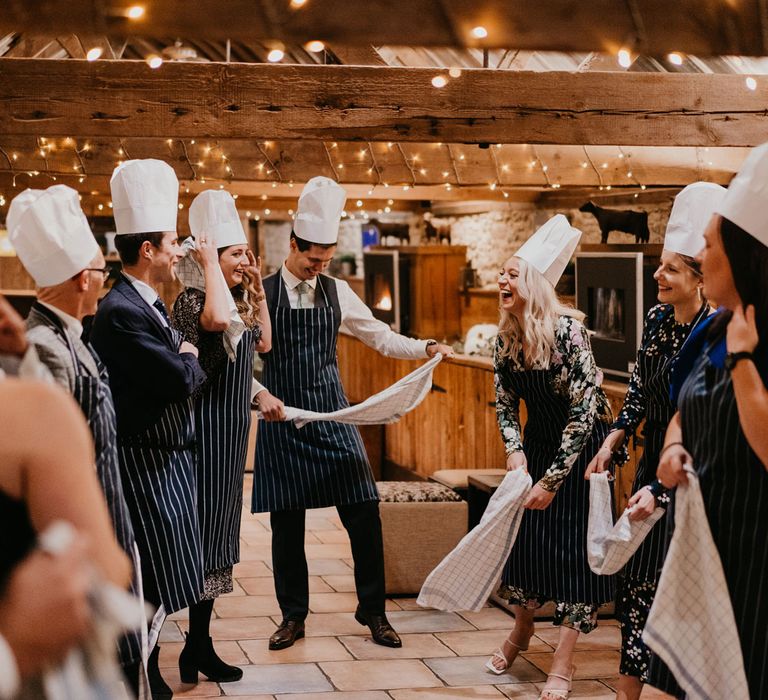 Groom and wedding guests in striped aprons and chefs hats stand in barn for rustic wedding reception