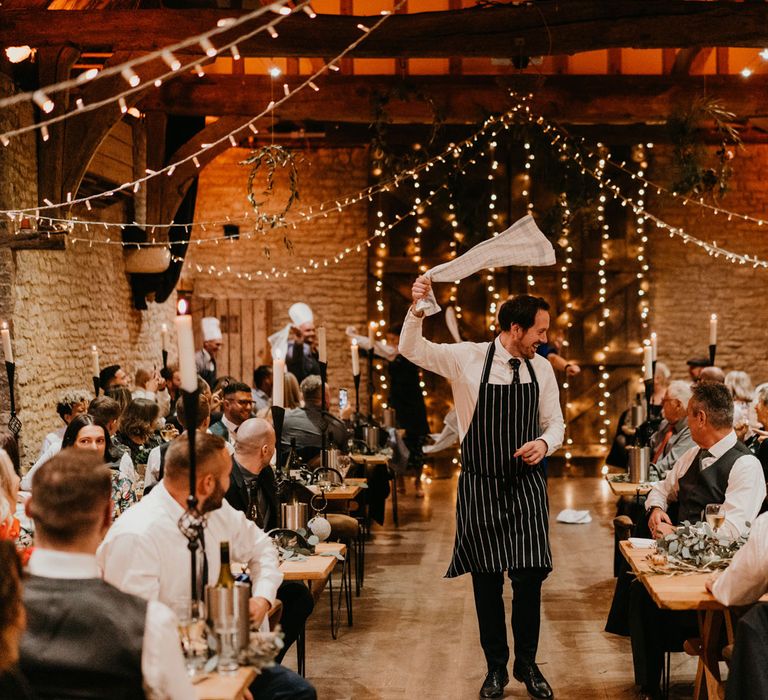 Server in striped apron swings white tea towel around as he walks down the middle of rustic wedding reception at The Tythe Barn 