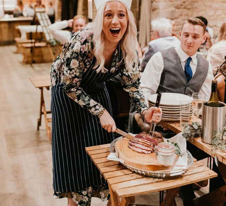 Guest in striped apron and chefs hat laughs as she cuts up meat for a guests table during rustic barn wedding breakfast