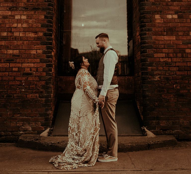 Boho bride in a lace wedding dress with long sleeves holding hands with her groom in a tweed waistcoat and Van trainers 