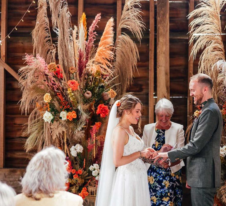 Bride in thin strap corset back wedding dress and veil exchanges rings with groom in green suit in front of rose and pampas grass floral installation during rustic barn wedding ceremony