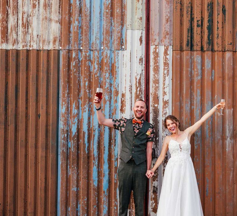 Bride in lace wedding dress holding glass of sparkling wine and groom in patterned short sleeve shirt, waistcoat and bow tie holding pint of ale raise their glasses