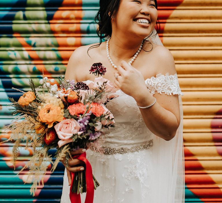 Laughing bride with pearl necklace and lace bardot wedding dress holding colourful wedding bouquet stands in front of street art by Scale wedding venue in Liverpool