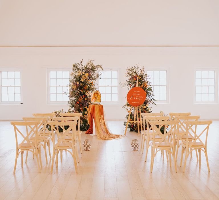 Golden wedding ceremony at Core Clapton Events wedding venue with wooden chairs, orange wedding sign and column flower arrangements 