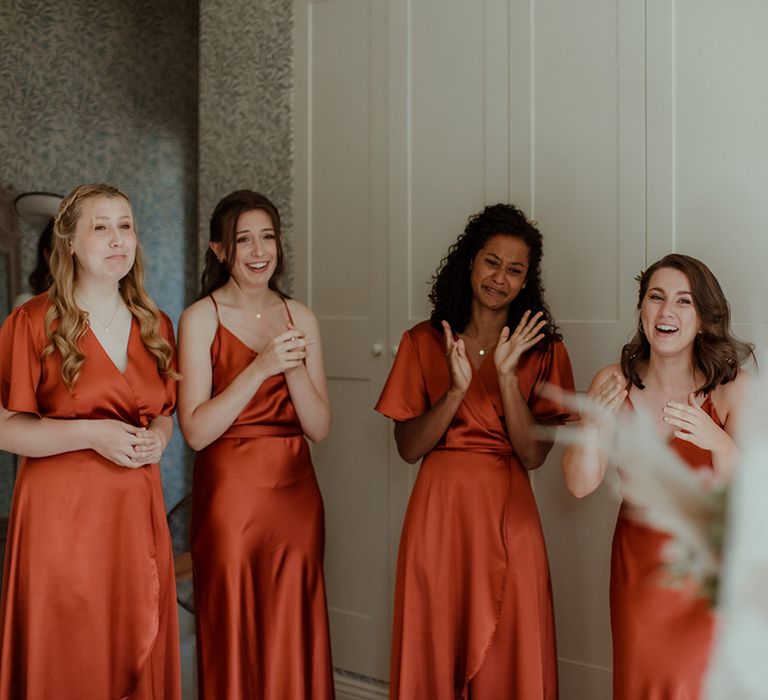 Bridesmaids in orange satin dresses in various styles cry and look in awe at the bride as she reveals her dress 