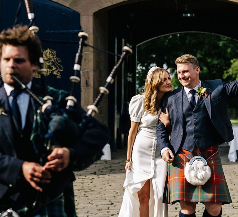 Bride in puff sleeve Bon Bride dress with pearl headband standing with groom in kilt and navy jacket with bagpipe player 