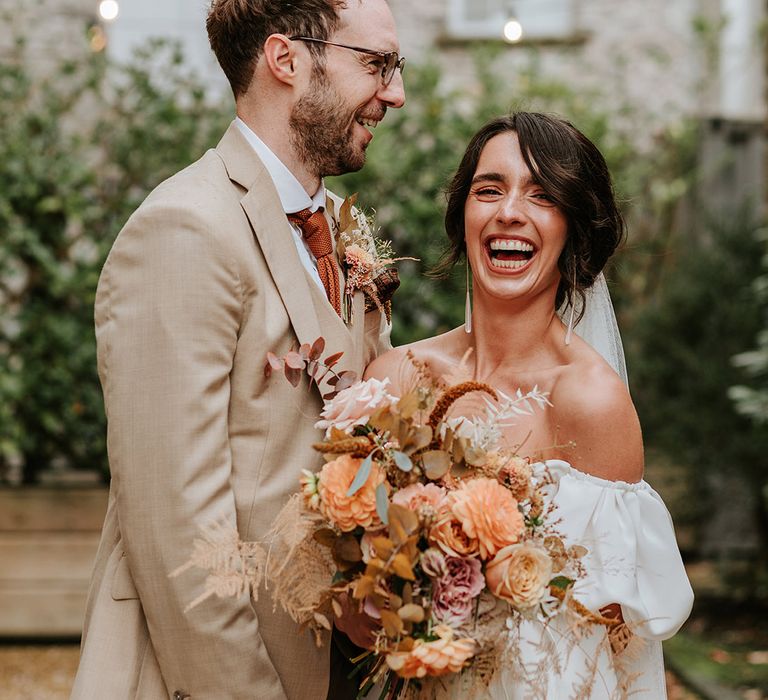 Laughing bride in puff sleeve wedding dress with groom in beige suit stand closely together