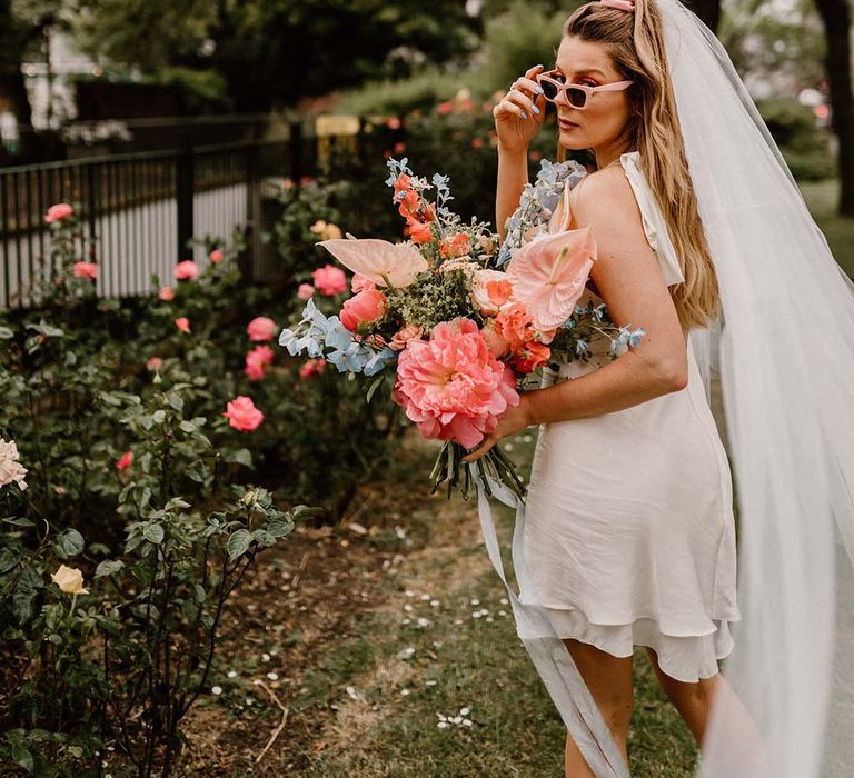 Stylish bride in a short wedding dress with retro sunglasses and long veil holding a pink and blue wedding bouquet 