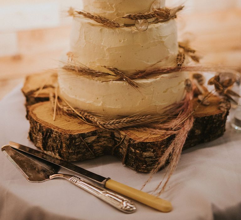 Three tier rustic looking white frosted cake with pieces of wheat on top of tree trunk cake stand 