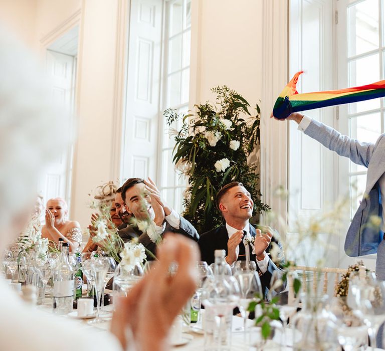 Groom's father in pale blue suit with patterned tie holds up rainbow scarf for pride at son's wedding