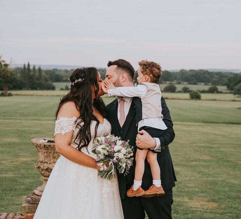 Young page boy pushes the brides and groom's faces as they lean in for a kiss