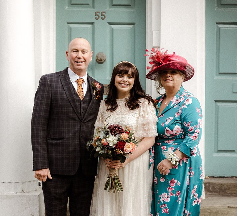 Bride holding rose wedding bouquet with autumnal coloured flowers poses with her parents