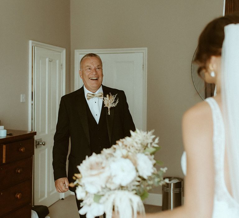 Father of the bride in black tie with gold bow tie smile widely as he gets first look at the bride