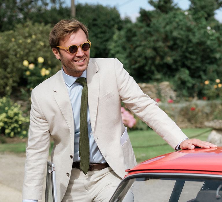 Groom in a sand coloured suit and sunglasses getting into his red car at his at home, tipi wedding 