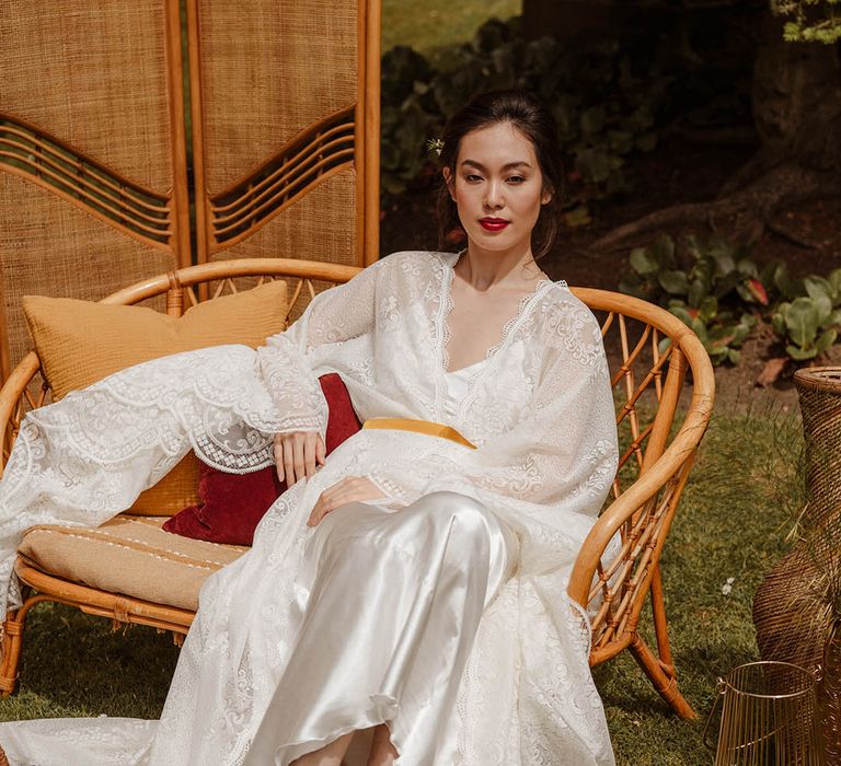 East Asian bride in a satin slip wedding dress with lace kimono style overlay sitting on a wicker chair at Japanese wedding shoot 