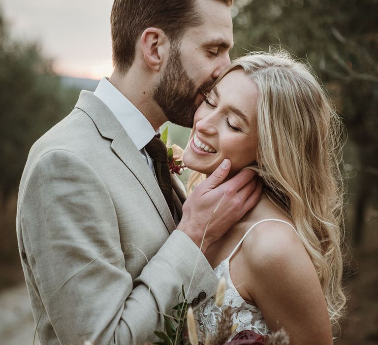 Groom kisses bride on the cheek as she smiles and holds colourful floral bouquet 