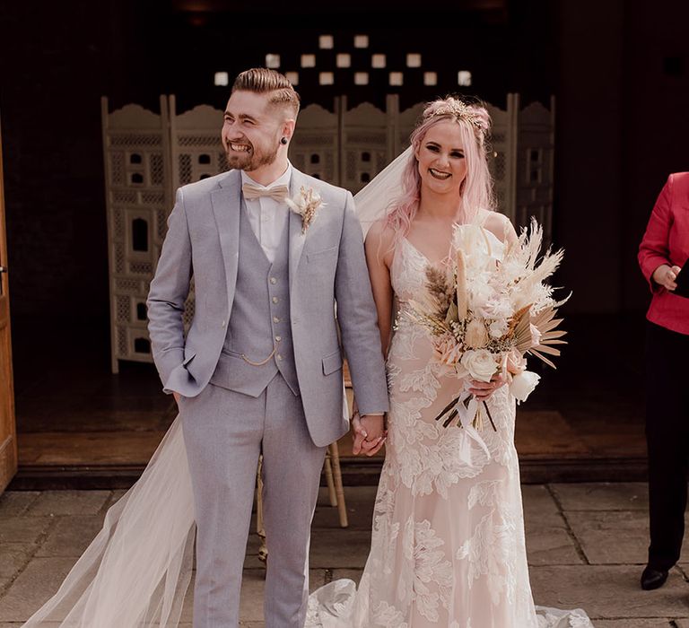 Bride with pink hair wearing Jane Hill boho fitted wedding gown with lace embellishment and floral bridal crown with floor-length veil carrying dried floral bouquet