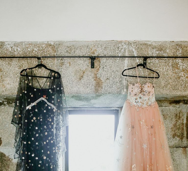 Bride's pink starry wedding dress and black celestial reception dress hang either side of window before wedding day