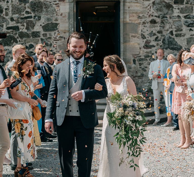 Bride wearing lace wedding dress and holding oversized bridal bouquet holds hands with her groom who wears three piece suit as confetti is thrown around them 