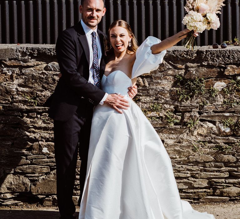 Bride in puff sleeve wedding dress with groom in a navy blue suit lifting up a pampas grass bouquet with pink roses and hydrangeas 