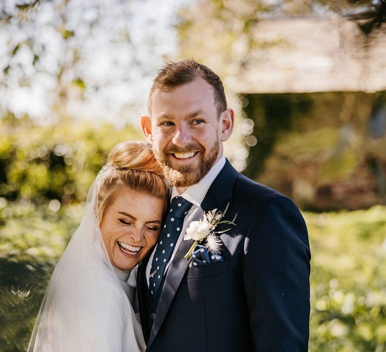 Bride in a high neck wedding dress laughs and rests her head on the groom's chest who wears a blur suit and polka dot tie 