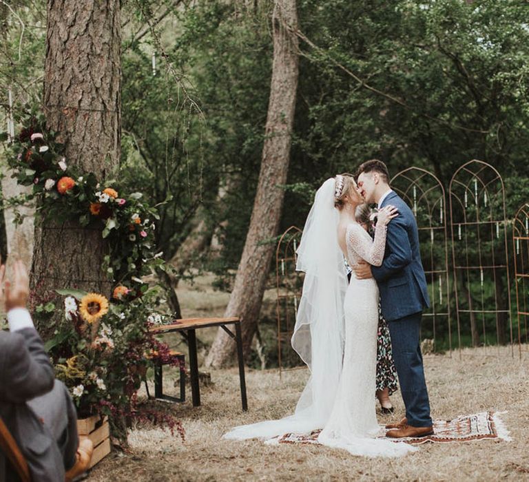 Bride and groom stand on a vintage rug as they share their first kiss as a married couple at their outdoor woodland ceremony 