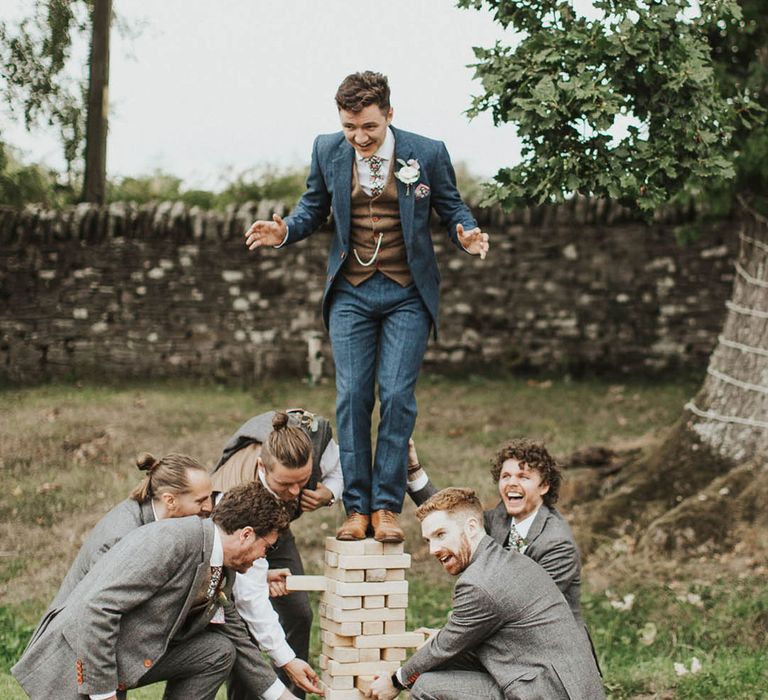 Groom in blue suit stands on top of jenga blocks as the groomsmen play 