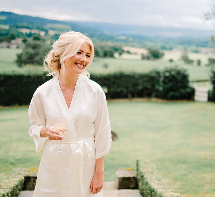 Blonde bride wears her hair pulled back into loose hairstyle as she holds a glass of champagne on the morning of her wedding day