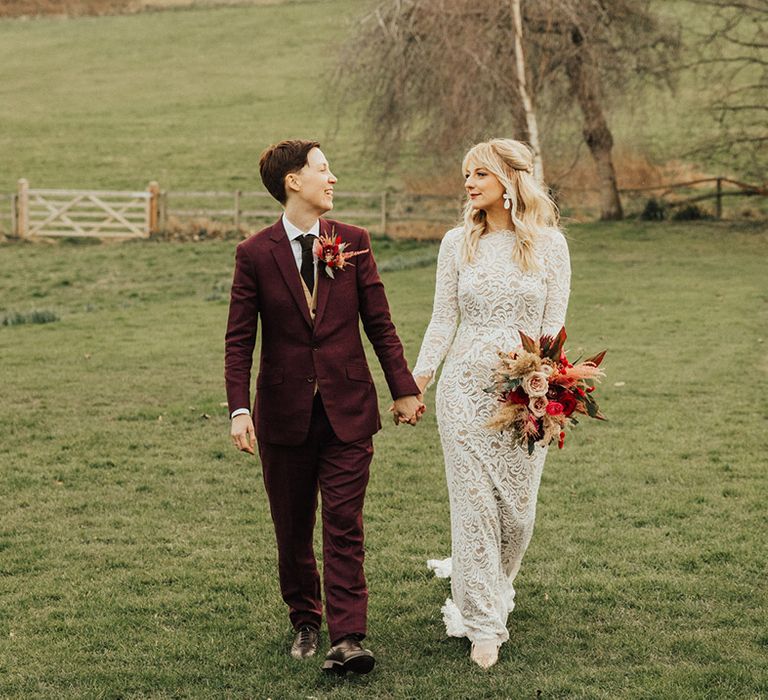 Bride in a high neck lace wedding dress walks holding hands with her partner in a burgundy suit 