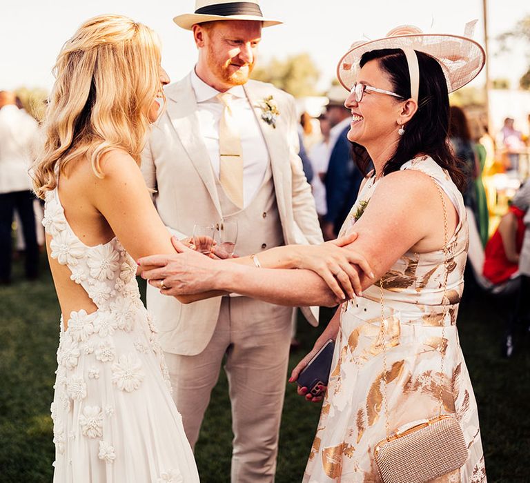 Bride in floral wedding dress and groom in cream suit and hat greets a wedding guest 