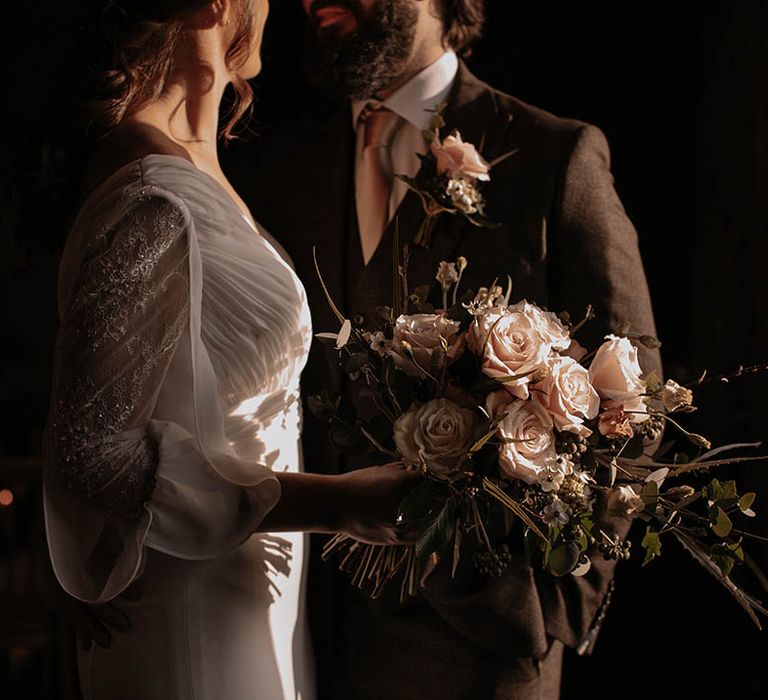 Bride in a sheer long sleeve wedding dress holding a pink rose winter wedding bouquet with the groom in a chocolate brown suit 