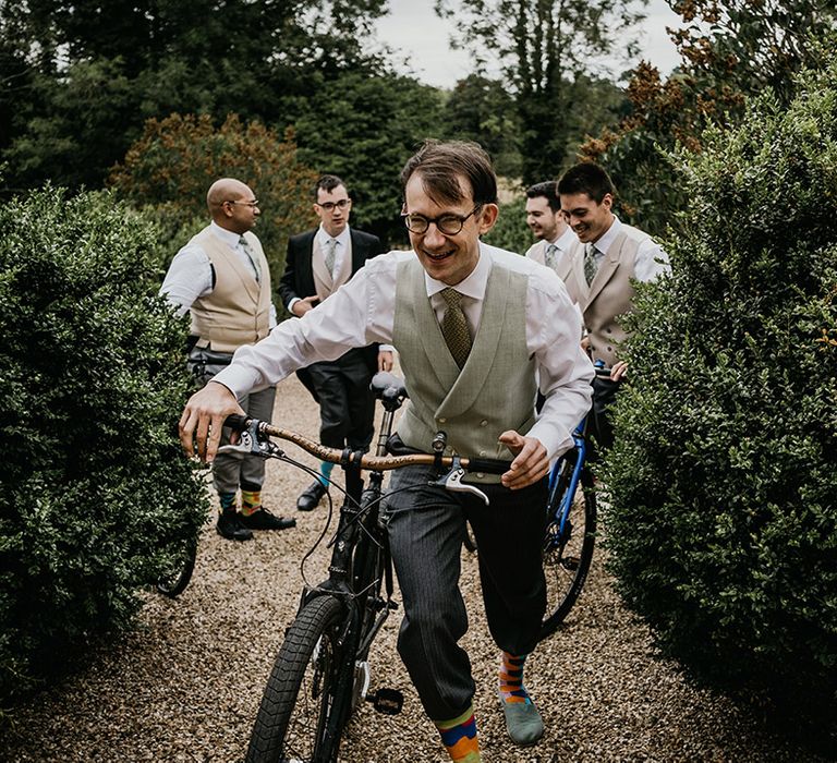 Groom in light coloured waistcoat wheeling his bike (his wedding transport) through the grounds of Kelmarsh Hall with groomsmen