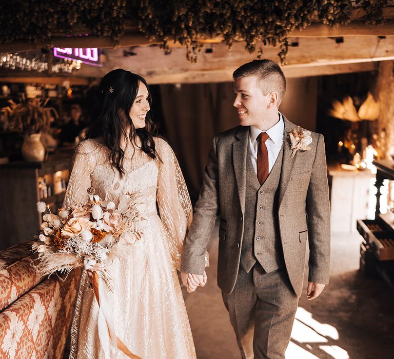 Bride in gold wedding dress holds dried floral bouquet and looks lovingly at her groom in three piece suit with burnt orange tie