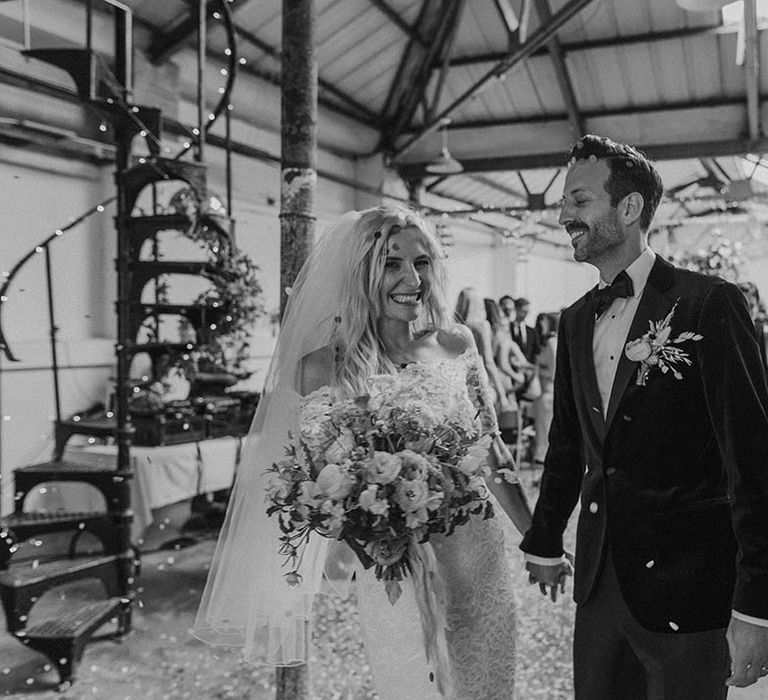 Bride in lace boho two piece with short veil and groom in black tux with bow tie and boutonniere standing amidst confetti at Hackney Studios